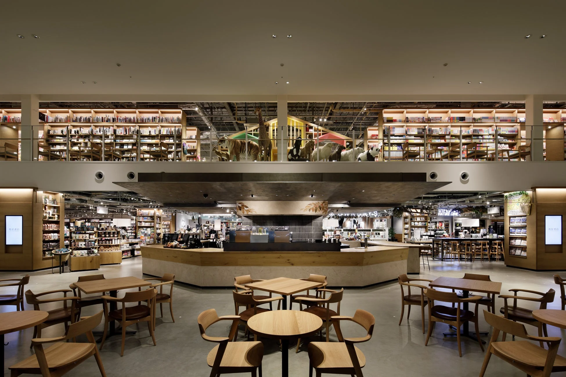 広々としたモダンな2階建ての書店とカフェの複合施設です。上階には本棚と動物のぬいぐるみが、下階にはカフェカウンターと多くの木製テーブルと椅子が配置されています。