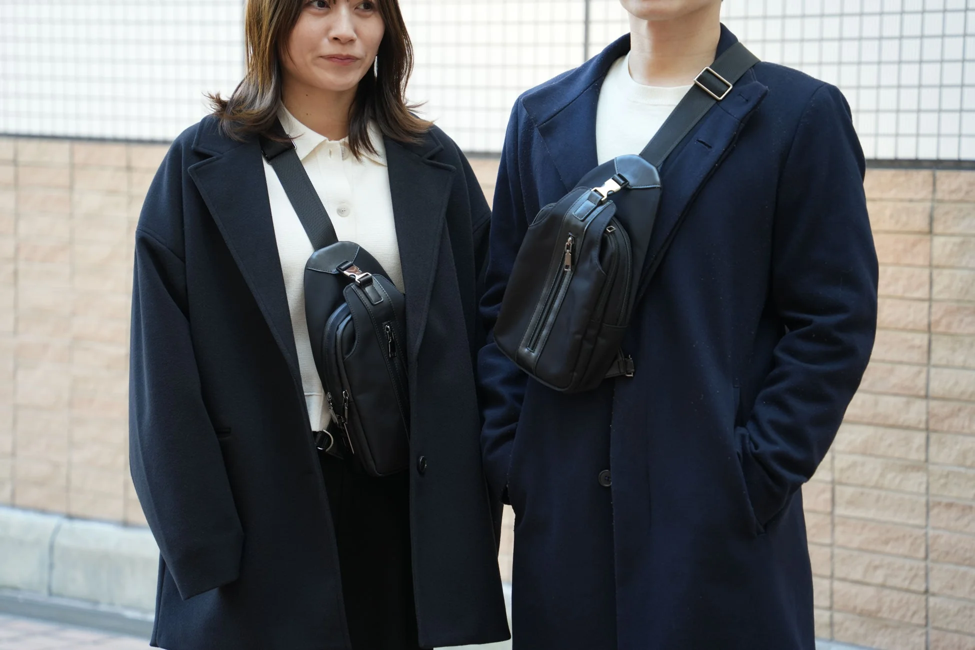 男女が黒いコートとスリングバッグを身につけて並んでいる様子。お揃いのバッグが印象的で、冬のカジュアルファッションを示しています。街中で撮影された、落ち着いた色合いのコーディネートです。