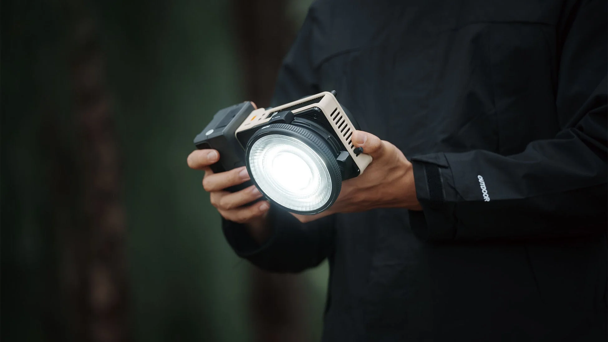 屋外で黒いジャケットを着た人物が、明るく発光する撮影用LEDライトを手に持っている様子を捉えた画像です。
