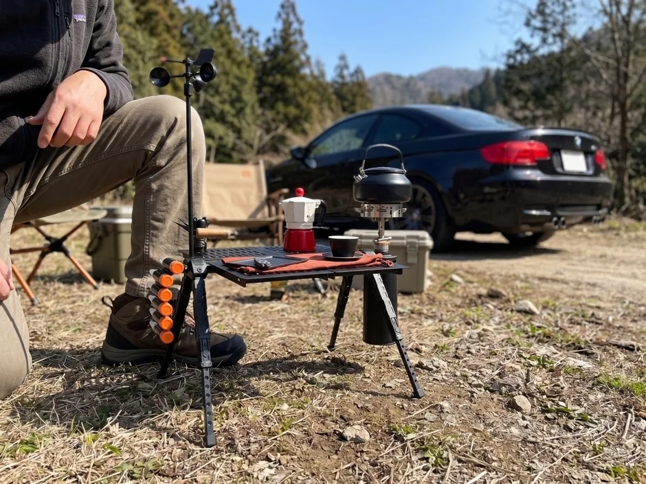 自然の中でポータブルテーブルを囲んでコーヒーを楽しむアウトドアシーン。黒い車が背景に停まり、風速計やキャンプチェアなどのギアが並べられている。