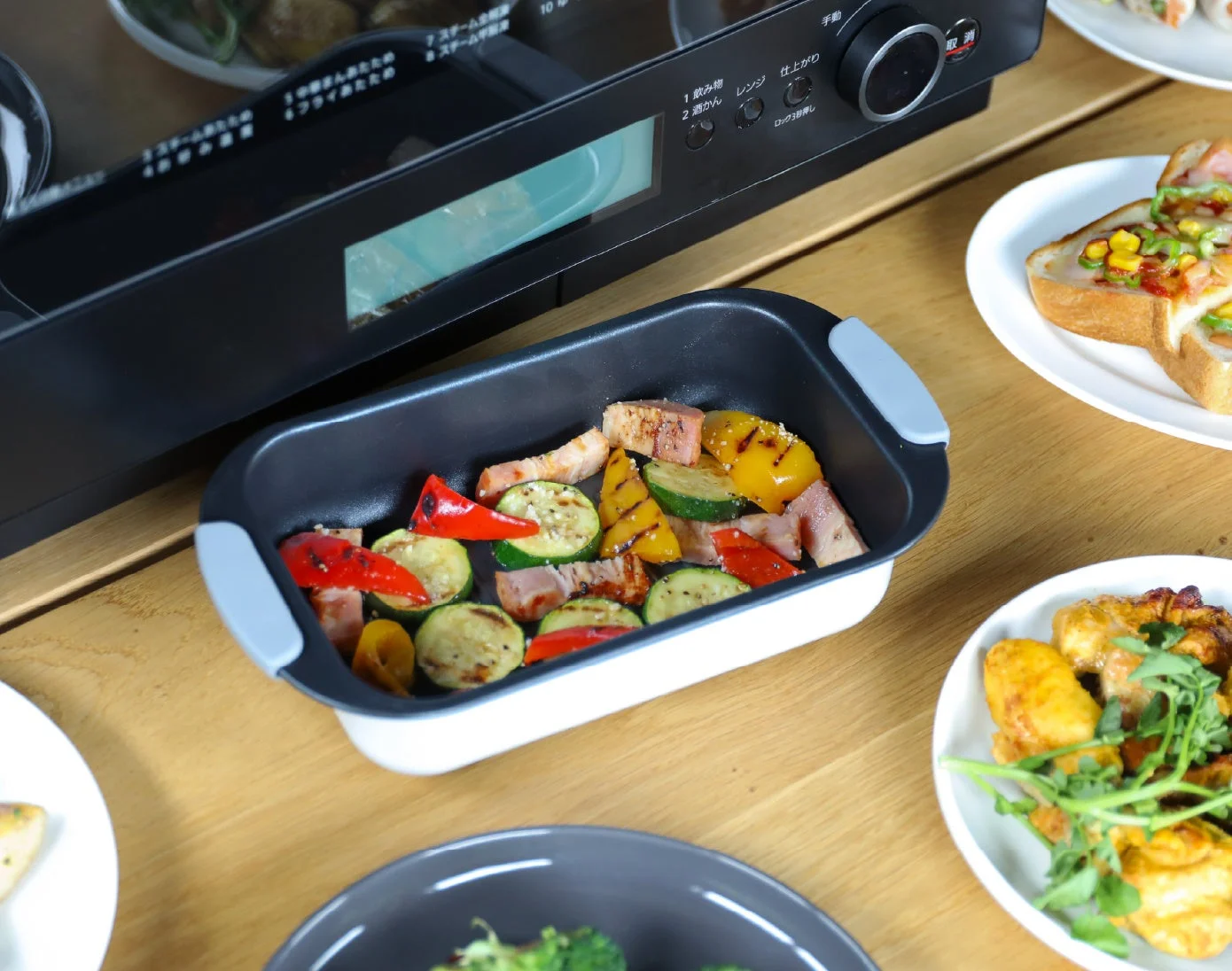 オーブンレンジの前に、グリル調理された色とりどりの野菜とベーコンが入った調理容器が置かれた食卓。他にもトーストやチキン料理が並び、複数の食事が準備されている様子が写っている。