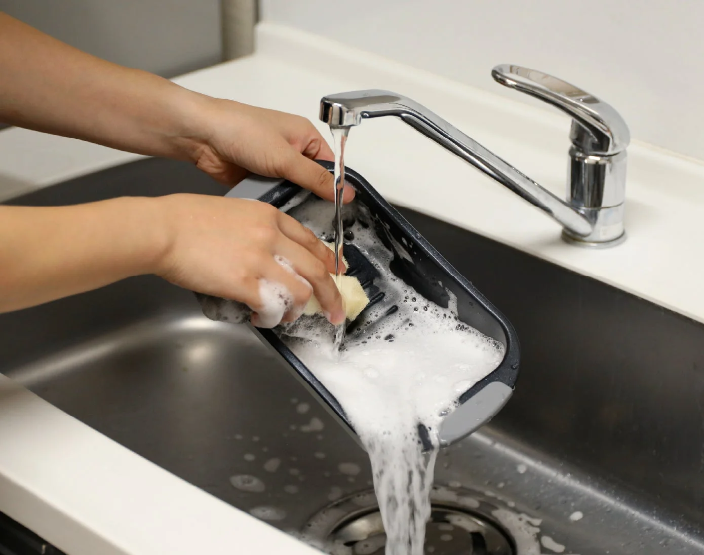 キッチンのシンクで、両手を使ってスポンジで容器を洗っている様子です。蛇口から水が勢いよく流れ出ており、容器の中には洗剤の泡がたっぷり立っています。清潔な状態にするための家事の一場面です。