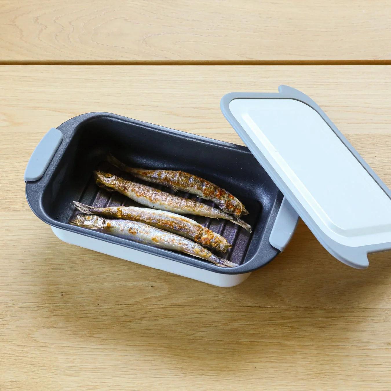 木目のテーブルに置かれた調理器具の中に、きつね色に焼かれたししゃもが数匹並べられています。調理器具の蓋は少し開いており、手軽に魚料理が楽しめる様子が伝わります。