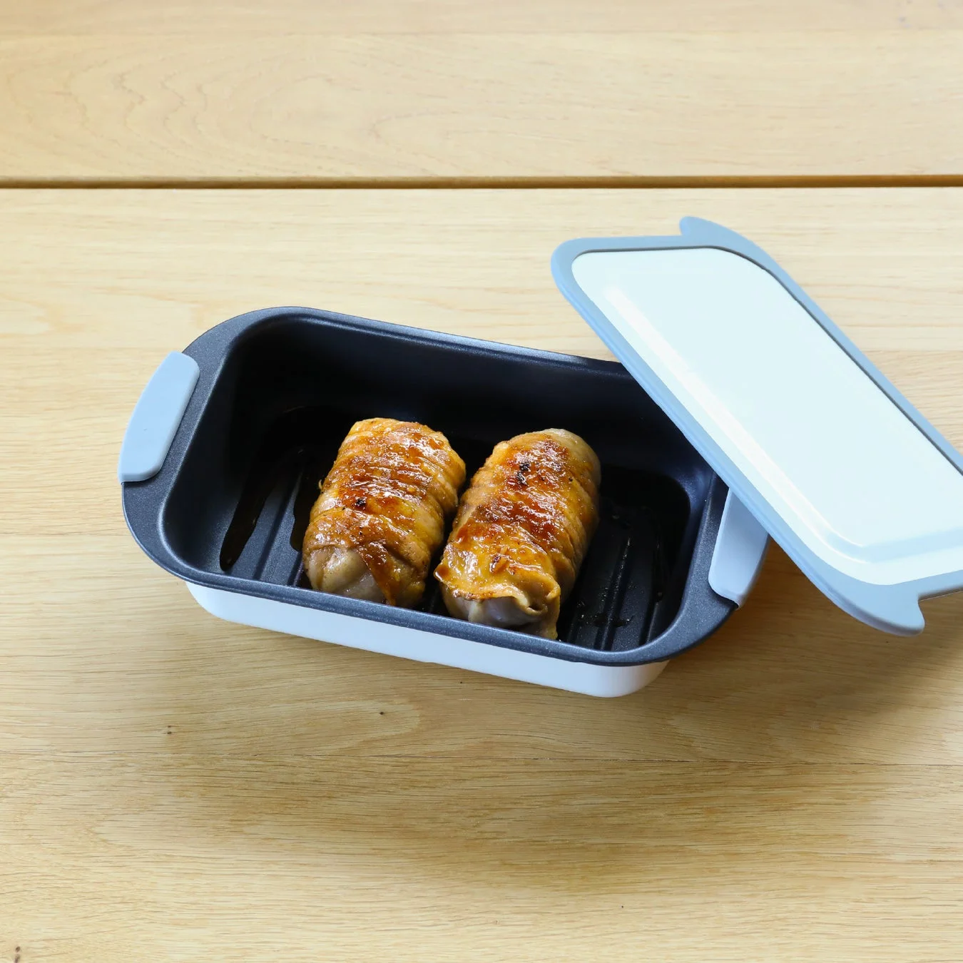 木製のテーブルの上に置かれた調理容器に、甘辛いタレで照り焼きにされたような肉巻きが2つ入っています。容器の蓋は開けられた状態で横に置かれています。