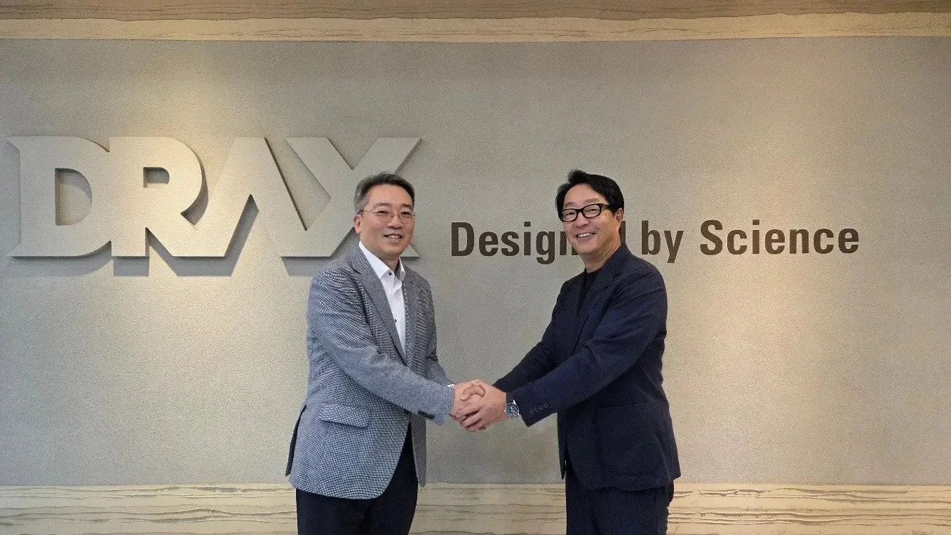 「DRAX Design by Science」のロゴがあるオフィスで、二人の男性が笑顔で握手をしているビジネスシーン。