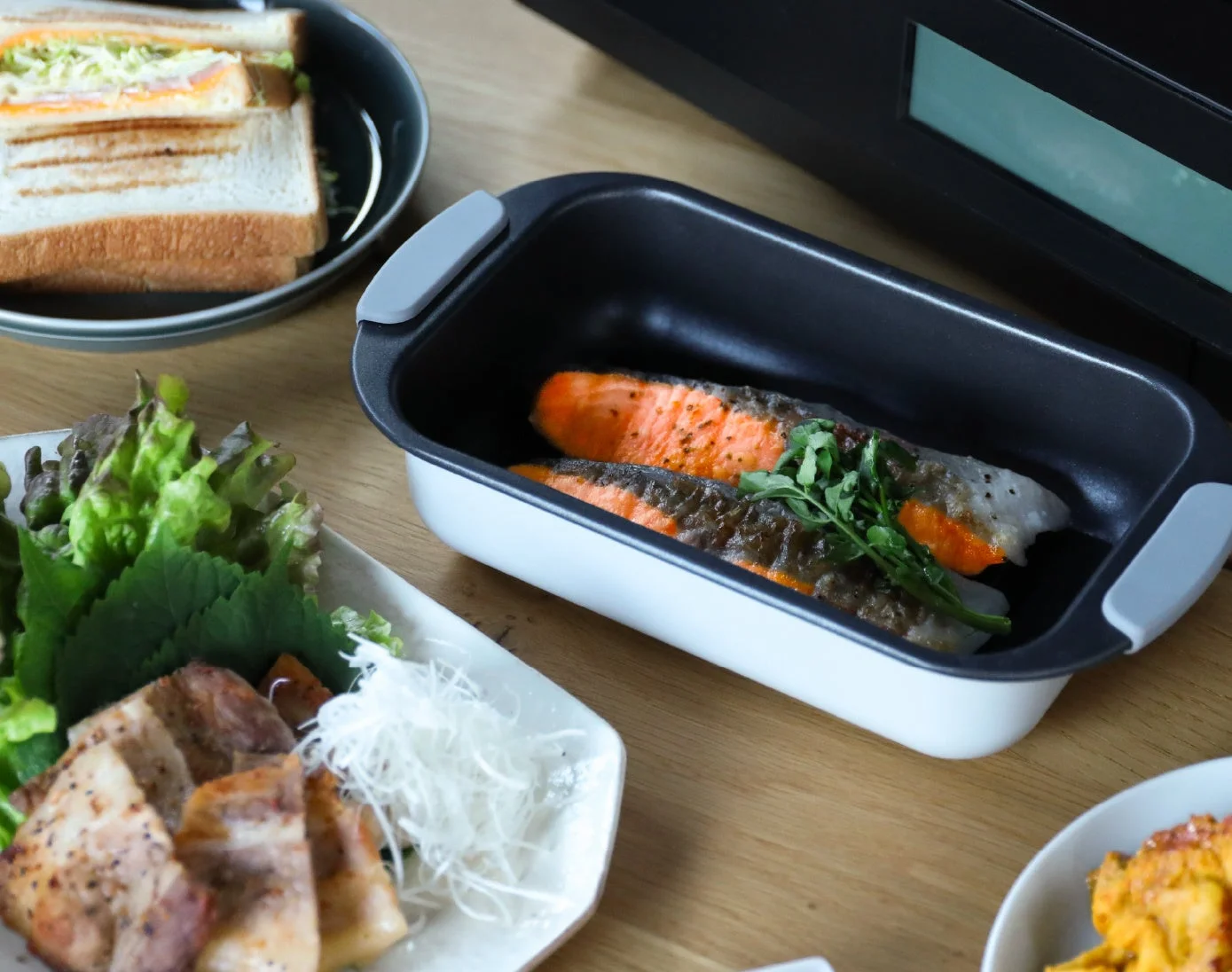 木目のテーブルに、焼き鮭、サンドイッチ、肉料理、野菜など様々な品が並べられた食事の風景です。バランスの取れた家庭料理の食卓の様子がうかがえます。