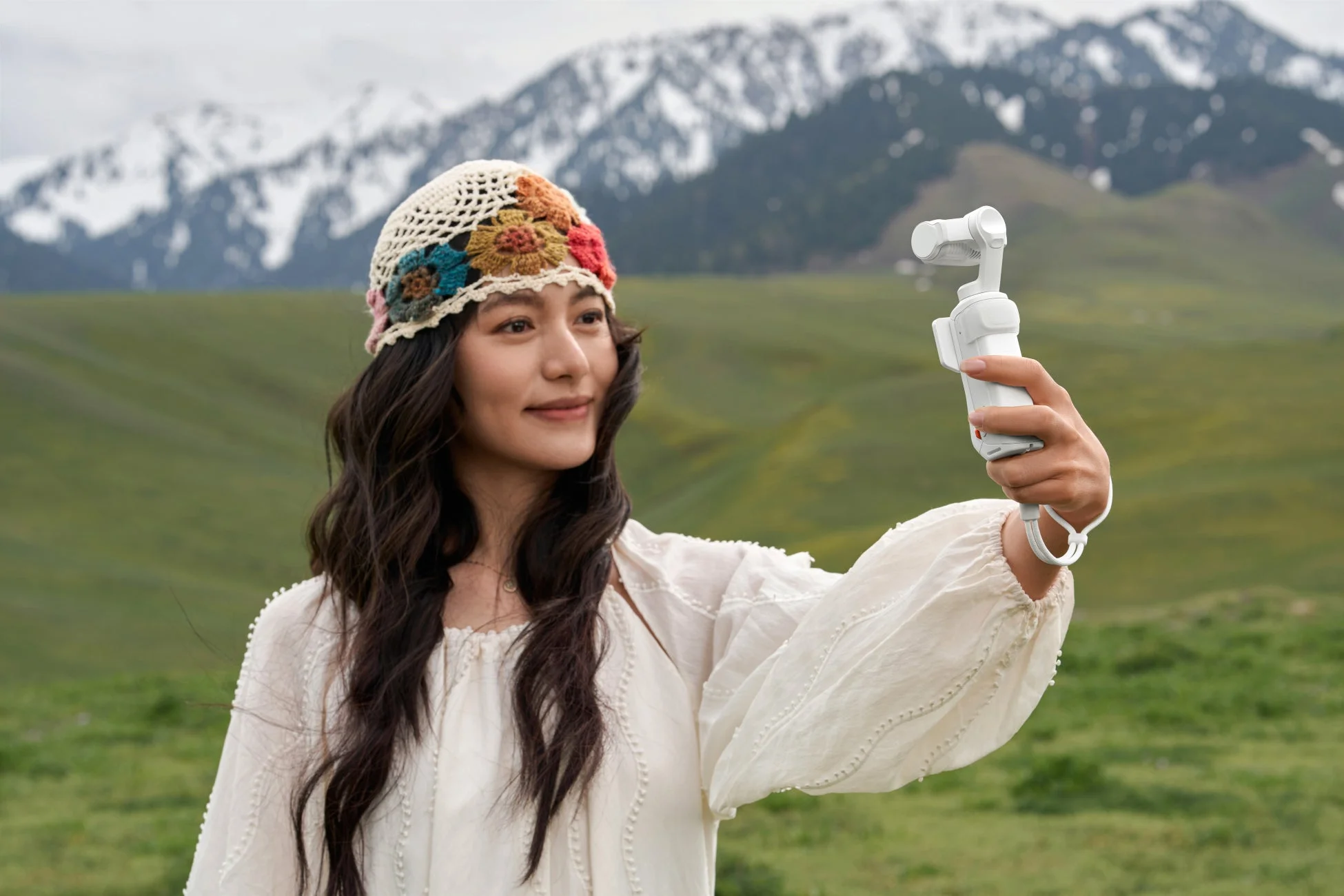 雪山と緑豊かな草原を背景に、民族調の帽子をかぶった女性が笑顔で白いカメラデバイスを手にしている。自然の中で撮影を楽しむ様子がうかがえる一枚です。