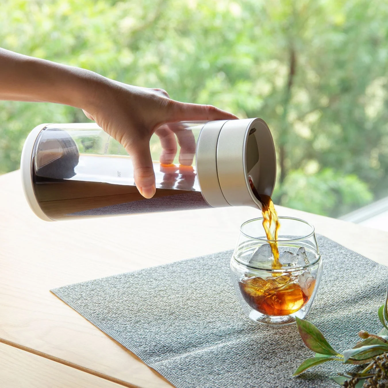 手がスタイリッシュなピッチャーからグラスに氷入りのコールドブリューコーヒーを注いでいる様子
