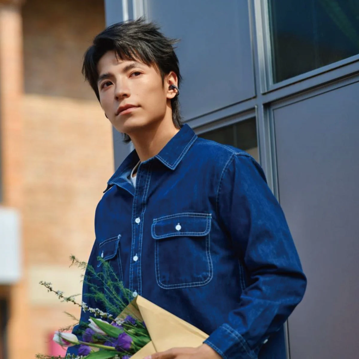 青いデニムシャツを着た若いアジア人男性が、耳にイヤホンをつけ、紫色の花束を抱えて立っている。