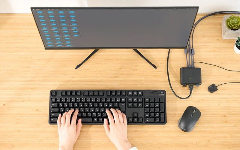 木目調のデスクに置かれたPC作業環境の俯瞰画像です。黒いキーボードを操作する両手、大型モニター、マウス、KVMスイッチが見られ、効率的なデスクワークの様子がうかがえます。