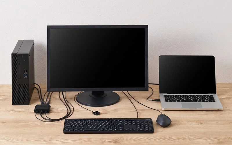 デスクトップPCとノートPCがKVMスイッチを介して一台のモニター、キーボード、マウスに接続されたPC環境が木製のデスク上に配置されています。複数のPCで周辺機器を共有するセットアップです。
