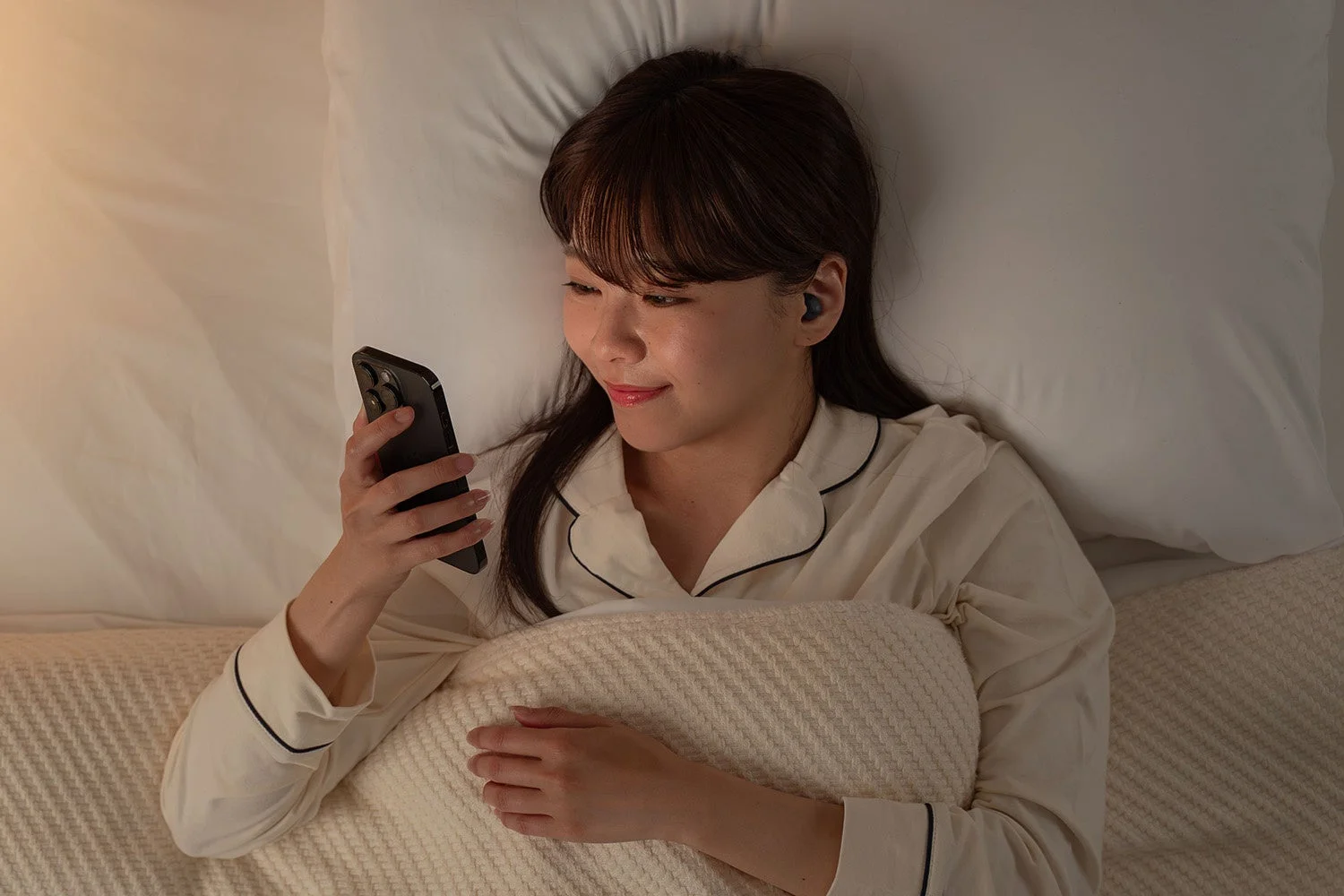 ベッドでパジャマ姿の女性が、イヤホンを装着してスマートフォンを操作しています