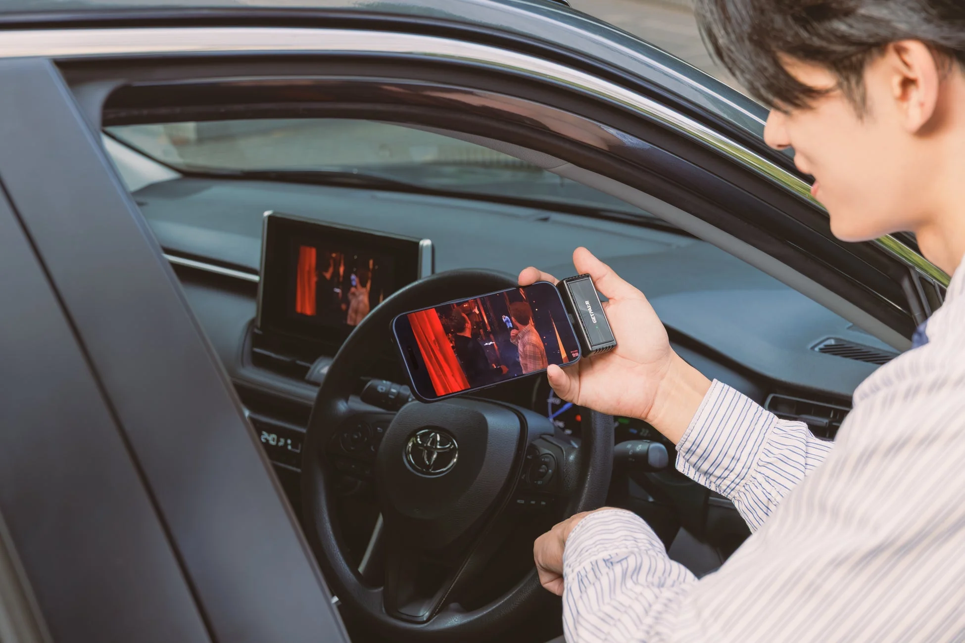 車内で男性がスマートフォンに接続されたデバイスを手に動画を視聴している様子です。車のインフォテインメントシステムにも同様の映像が映し出されています。