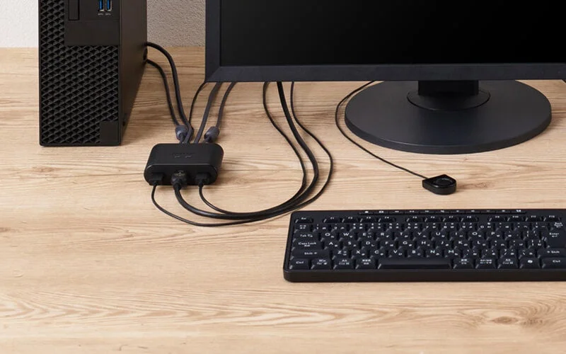 木目調のデスク上に、デスクトップPC、モニター、キーボードが配置され、KVMスイッチと多数のケーブルで接続されている作業環境の様子