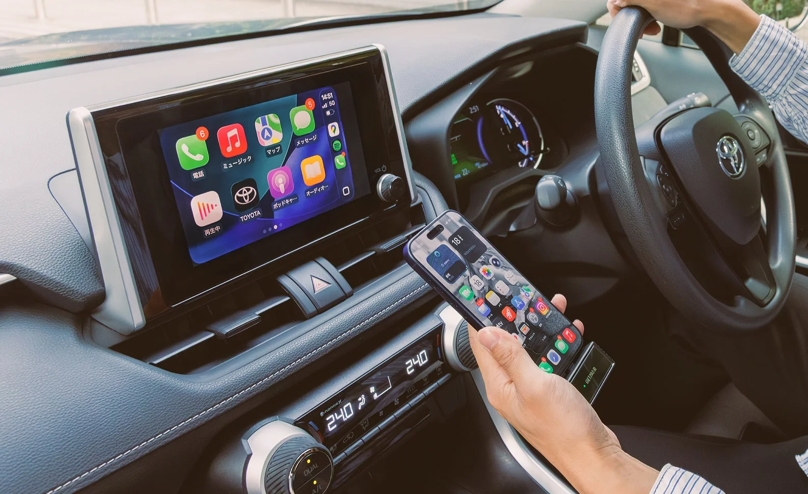 車内で運転席に座る人物がスマートフォンを持ち、車のディスプレイにApple CarPlayが表示されている様子。