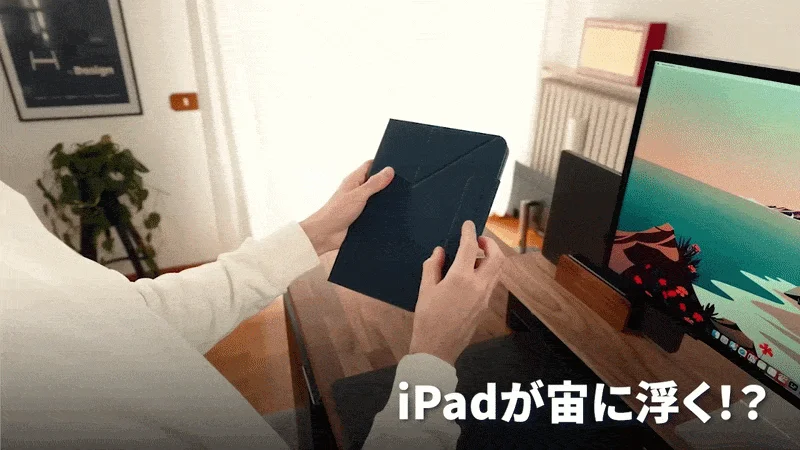一人の人物がデスクの前でタブレットケースを持っており、その手元には「iPadが宙に浮く!?」というテキストが表示されています。背景にはモニターや観葉植物が見え、整頓された部屋の一角での作業風景を捉えています。