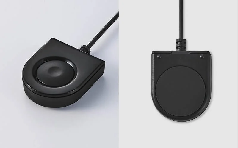黒いフットスイッチのような入力デバイスが、斜めと真上からの2つのアングルで写っています。中央に大きな丸いボタンまたはパッドがあり、シンプルなデザインです。