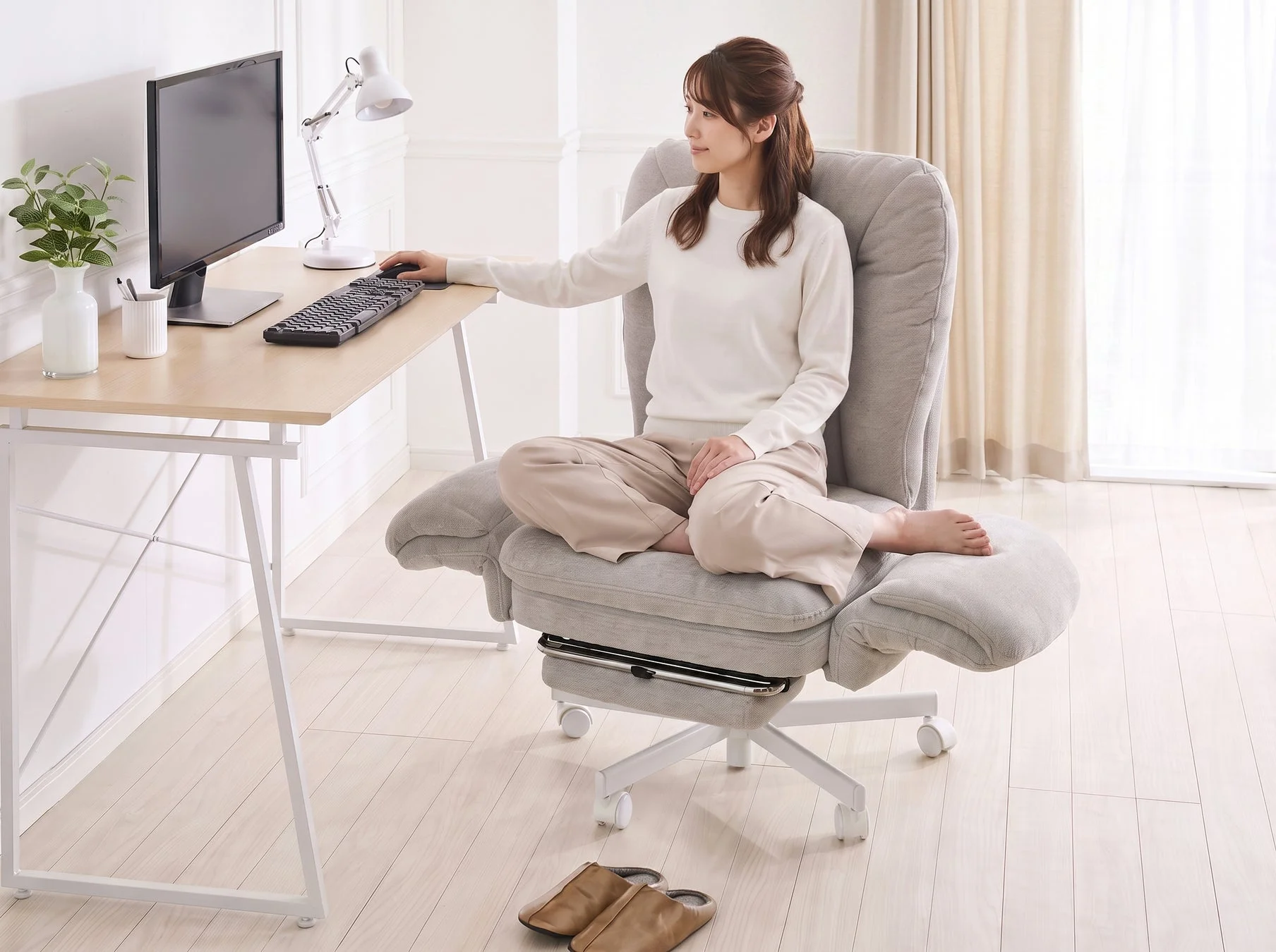 明るい部屋で、女性がグレーの快適なオフィスチェアに座り、木製のデスクでパソコンに向かっています。あぐらをかいてリラックスした姿勢