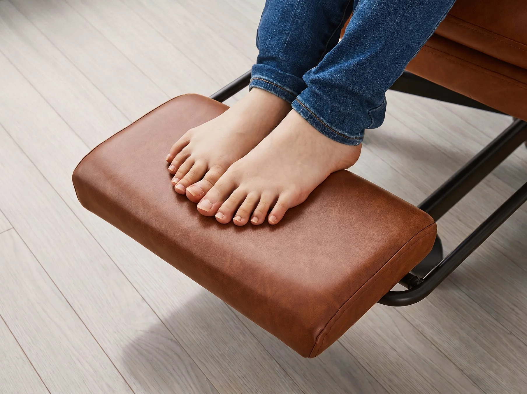 素足の人物が、茶色のレザー調のフットレストに足を乗せてリラックスしている様子