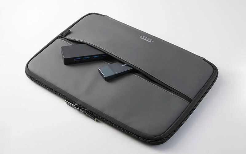 グレーのノートPC用スリーブケースの前面ポケットに、USBハブと小型のストレージデバイスが収納されている様子。