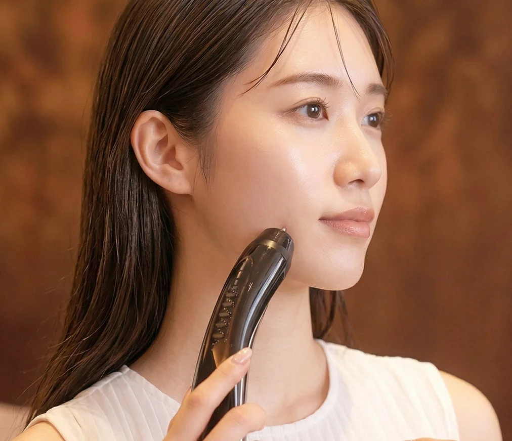 光沢のある肌の若い女性が、黒い美顔器を使って頬のケアをしている横顔のクローズアップ。美容やスキンケアのイメージです。