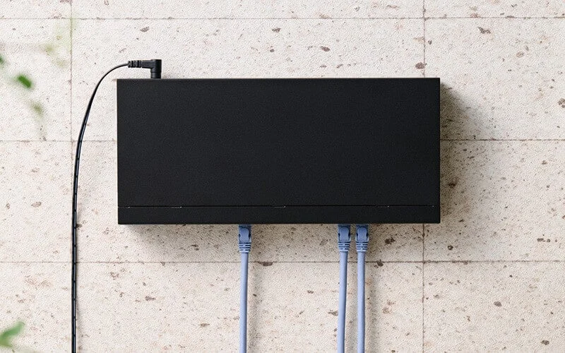 壁に設置された黒い長方形のネットワーク機器で、上部には電源ケーブル、下部には3本の水色のLANケーブルが接続されています。