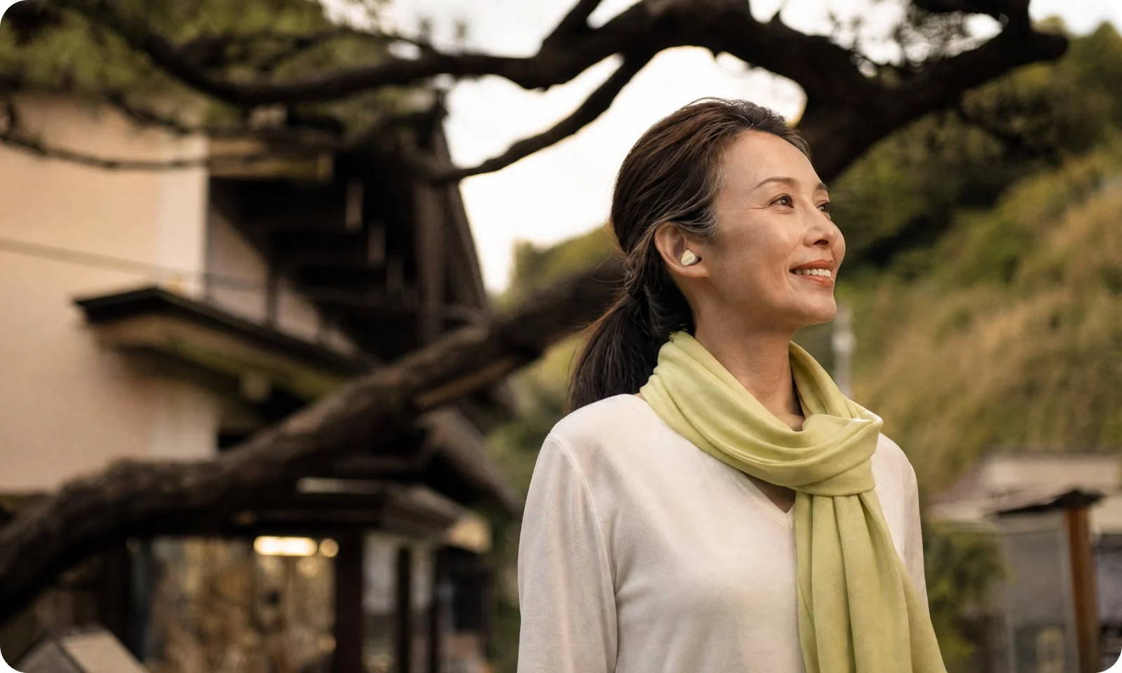 イヤホンとスカーフを身につけた中高年の女性が、伝統的な日本家屋と自然を背景に、屋外で穏やかに微笑んでいます。リラックスした雰囲気が伝わる一枚です。