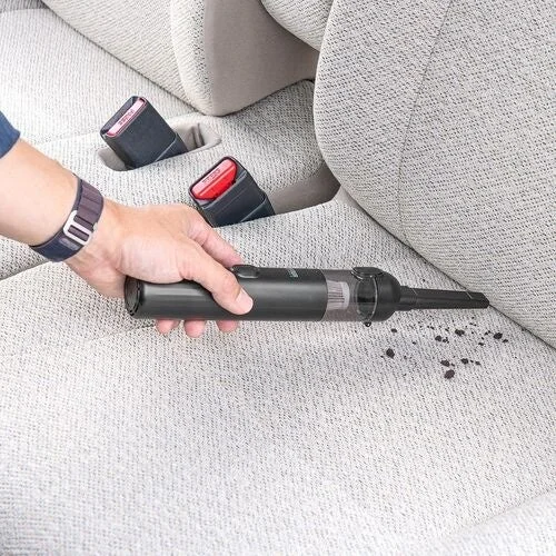 車内で、人がコンパクトなハンディクリーナーを使って車のシートに落ちたゴミを掃除している様子です。シートベルトも写っており、車内の清掃風景が分かります。