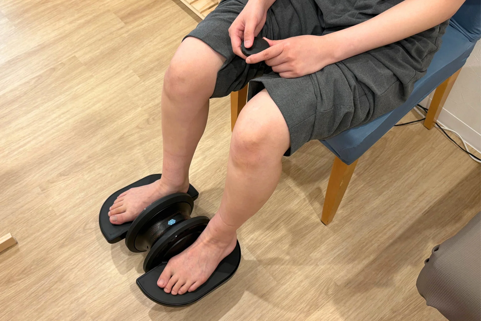 椅子に座った人物が、黒いフットマッサージャーに裸足を乗せて使用している様子です。室内でリラックスしながら足のケアをしている場面を捉えています。