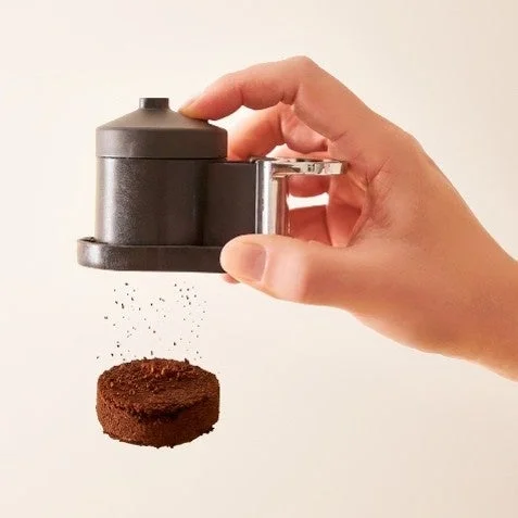 手が黒いコーヒー粉ディスペンサーを操作し、そこから挽かれたコーヒー粉が落ち、丸い固まりを形成している様子