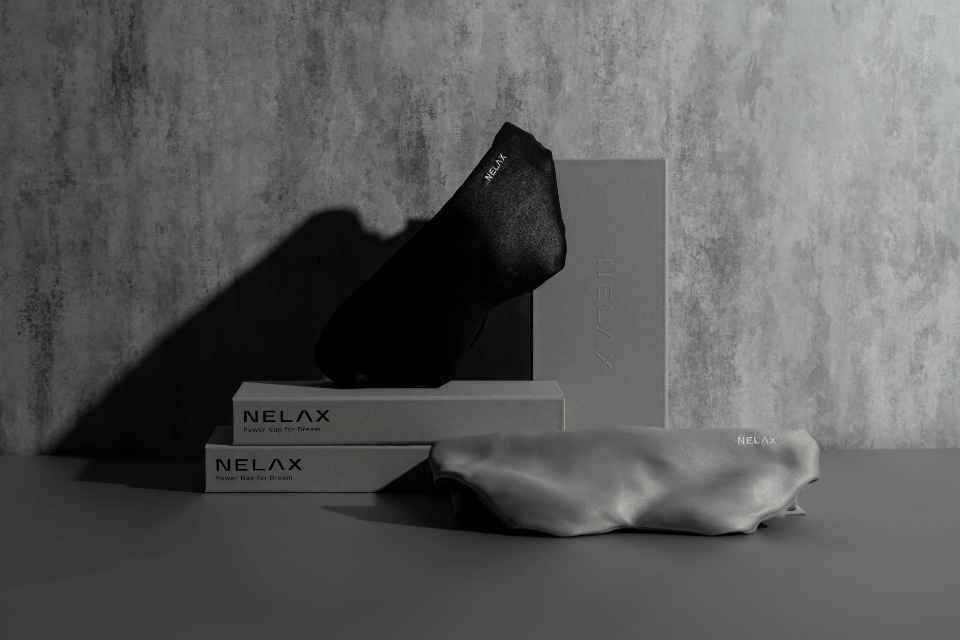 NELAXブランドのアイマスクとそのパッケージが写された商品写真です。黒とグレーのアイマスクが並べられ、コンクリート調の背景で、モノクロームかつミニマルな、落ち着いた雰囲気を演出しています。