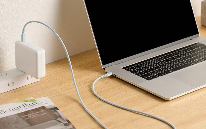 木製の机の上でノートパソコンが充電されており、電源タップと雑誌が写っています