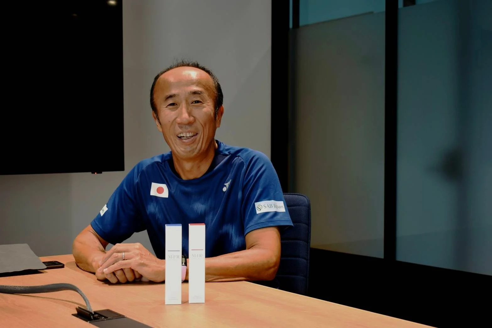 日本代表のスポーツウェアを着た男性が笑顔でテーブルに座り、手前には2つの白い商品パッケージが置かれている。インタビューかプロモーションの場面のようだ。