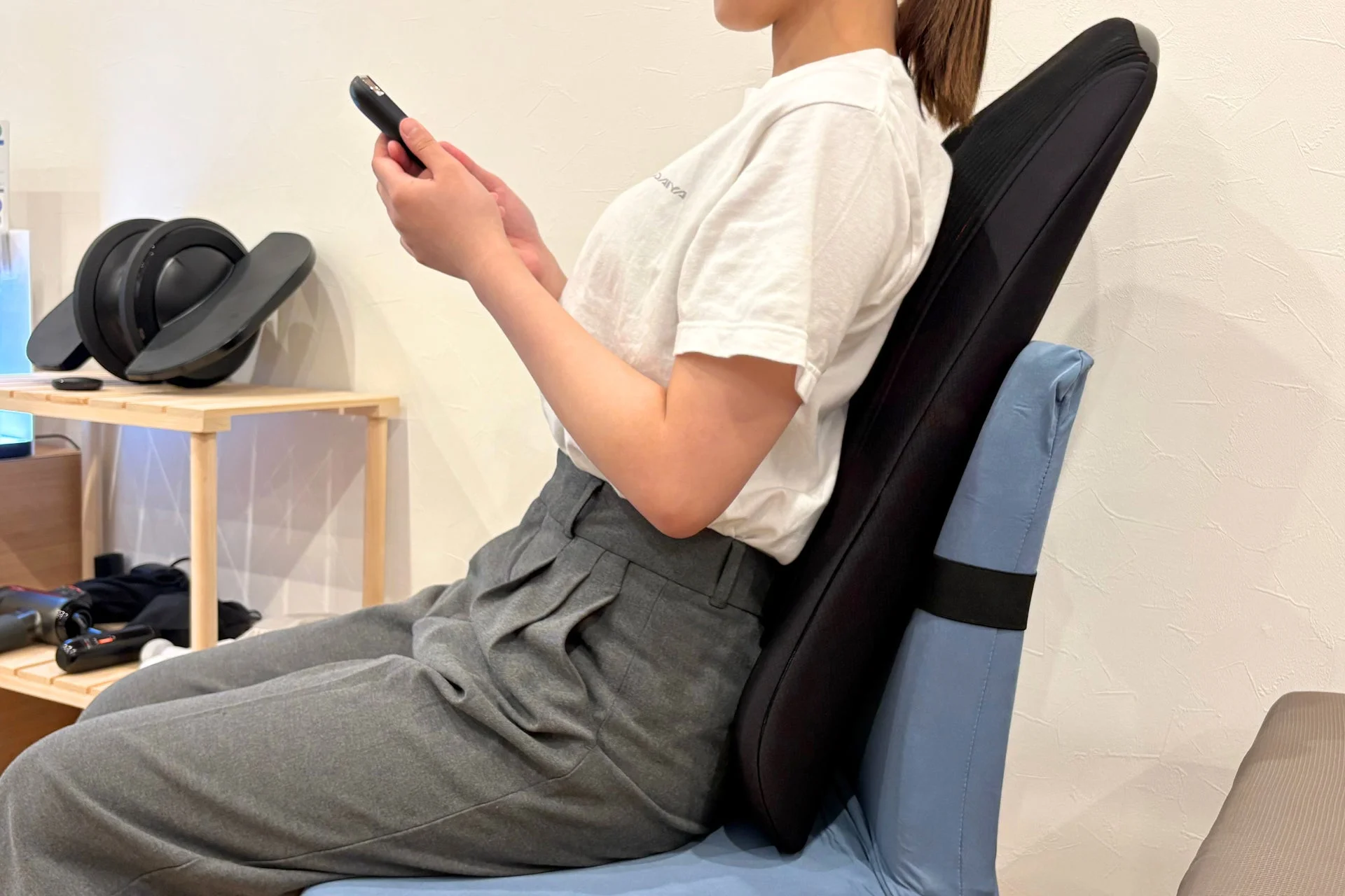女性が姿勢矯正クッションを背もたれにした椅子に座り、スマートフォンを操作しています。室内でリラックスした姿勢でモバイルデバイスを使用している様子が捉えられています。