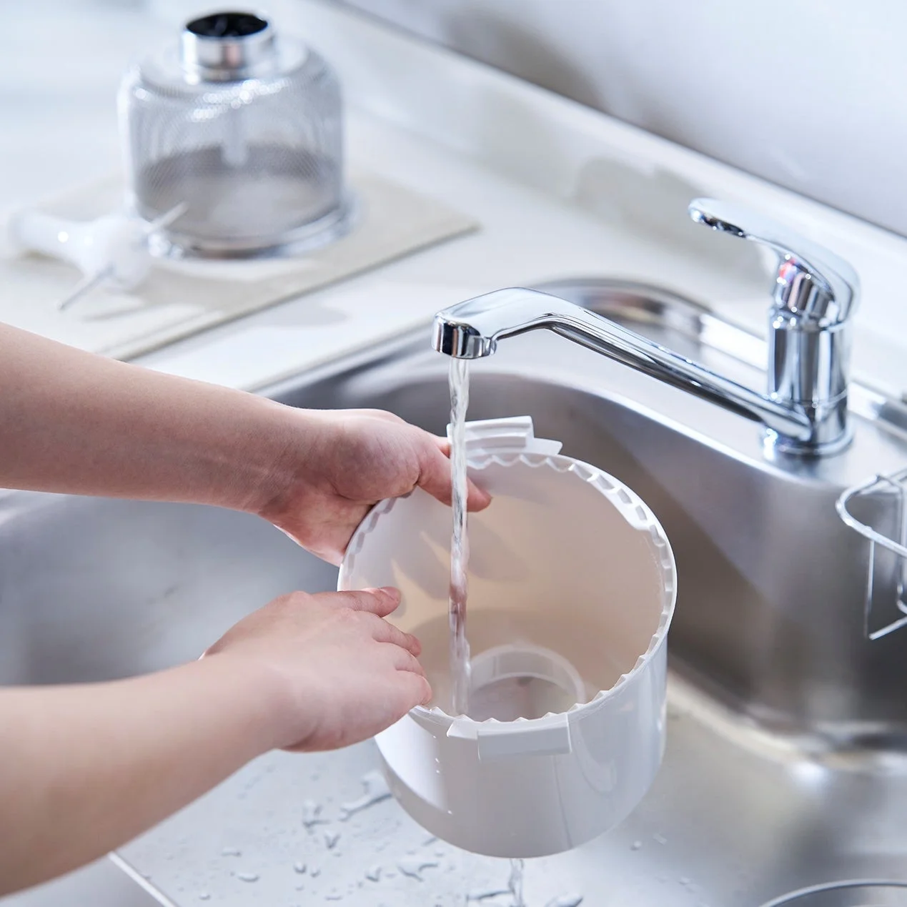 キッチンシンクで、人物が白い調理器具（おそらく炊飯器の内釜）を蛇口から出る水で洗っている様子を写した画像です。清潔感のある日常の家事風景です。