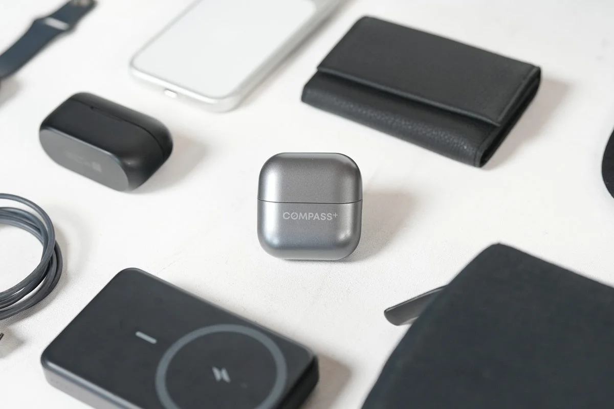 白い背景に、グレーのイヤホンケース「COMPASS+」を中心に、スマートフォン、財布、ワイヤレス充電器などの日用品やガジェットが整然と並べられている