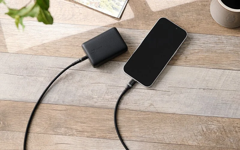 木目調のテーブルの上で、黒いモバイルバッテリーがスマートフォンに接続され充電している様子