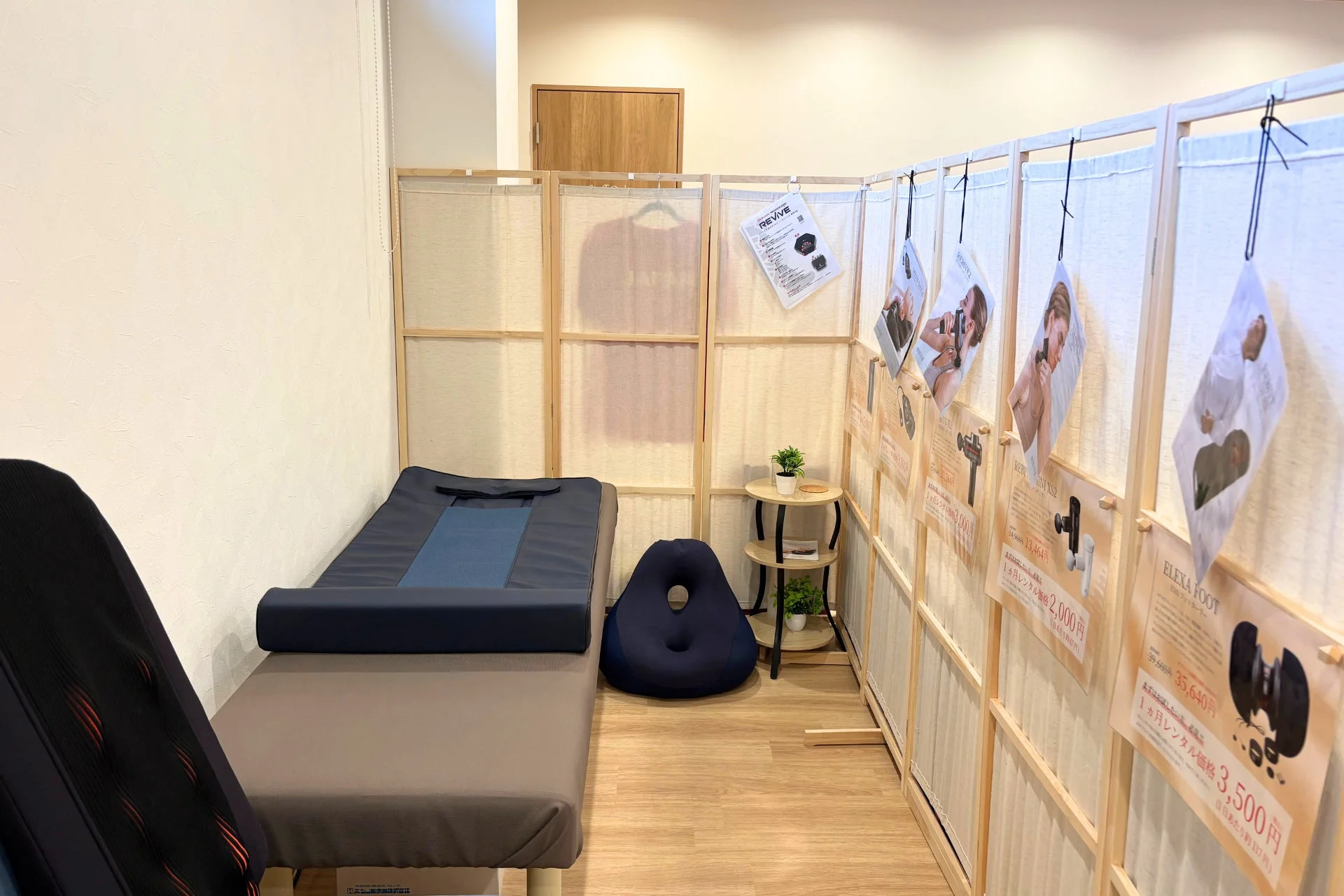 施術台が設置されたリラックス空間で、健康・美容機器のレンタルサービス「REVIVE」「ELE XA FOOT」「REB MINI XS2」の広告が多数掲示されている。各機器の1ヶ月レンタル価格や割引後の価格が案内されている。