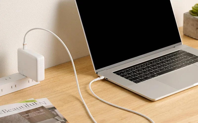 木製のデスクでノートパソコンが白い充電器とケーブルを使って電源タップから充電されている様子