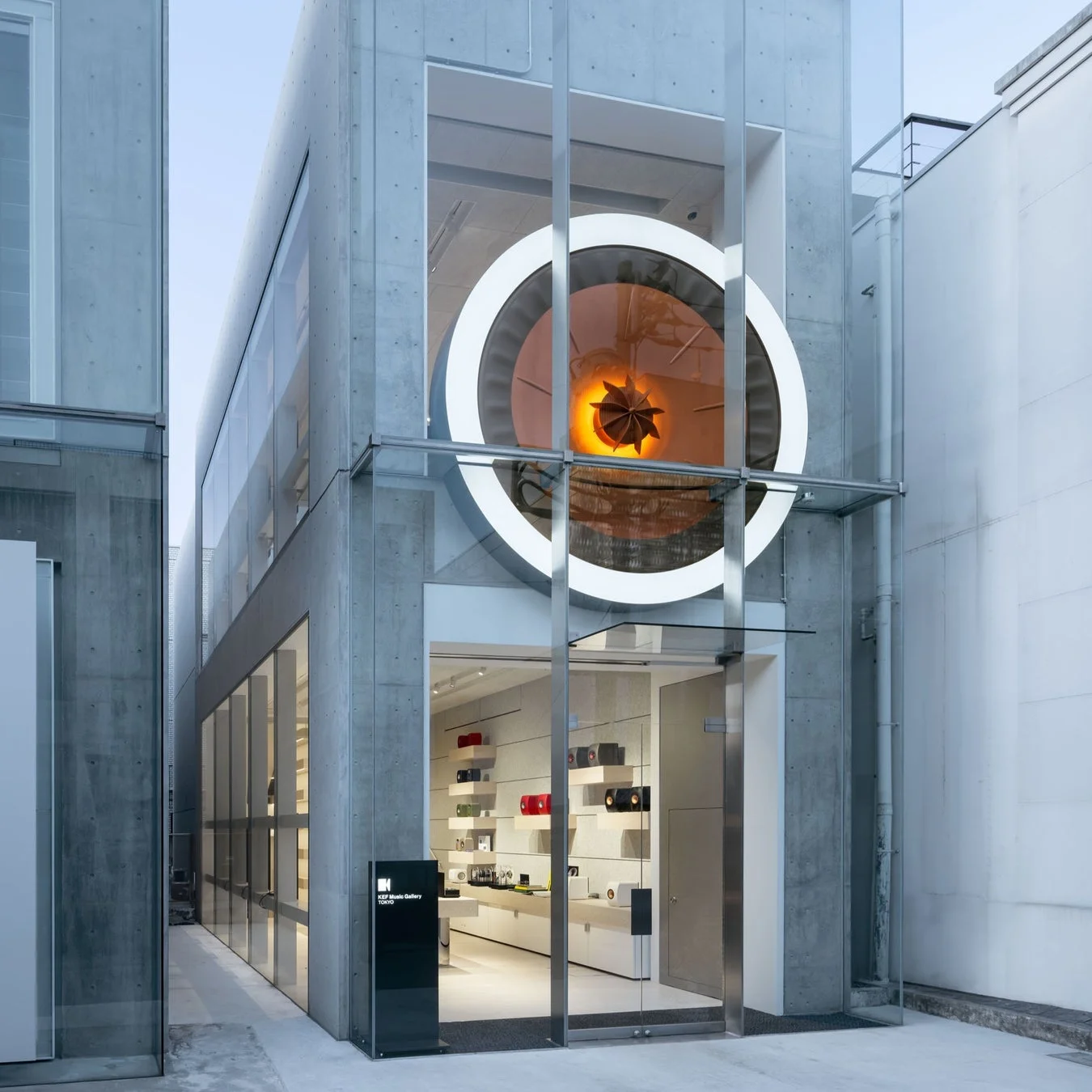 コンクリートとガラスを基調としたモダンな建築物で、上階の大きな丸窓から特徴的な照明付きのオブジェが見えるKEF Music Gallery TOKYOの店舗外観。