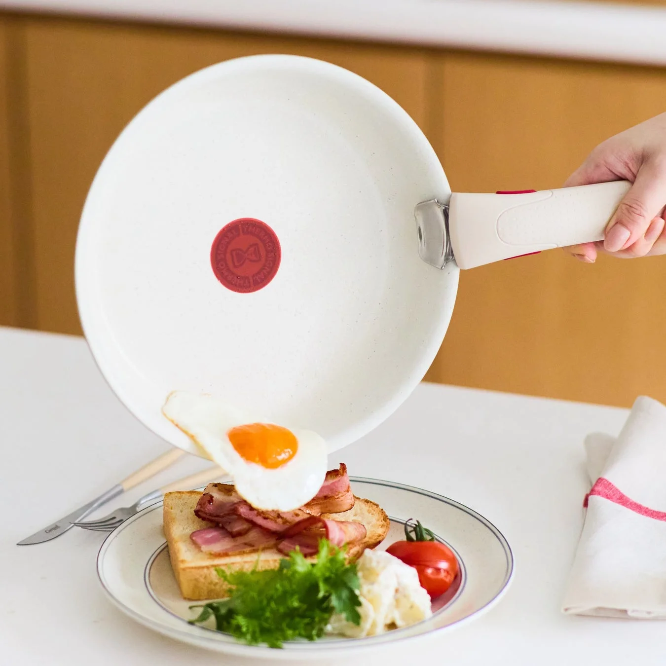 白いフライパンで焼いた目玉焼きが、ベーコンとトーストが乗ったお皿に盛り付けられている様子