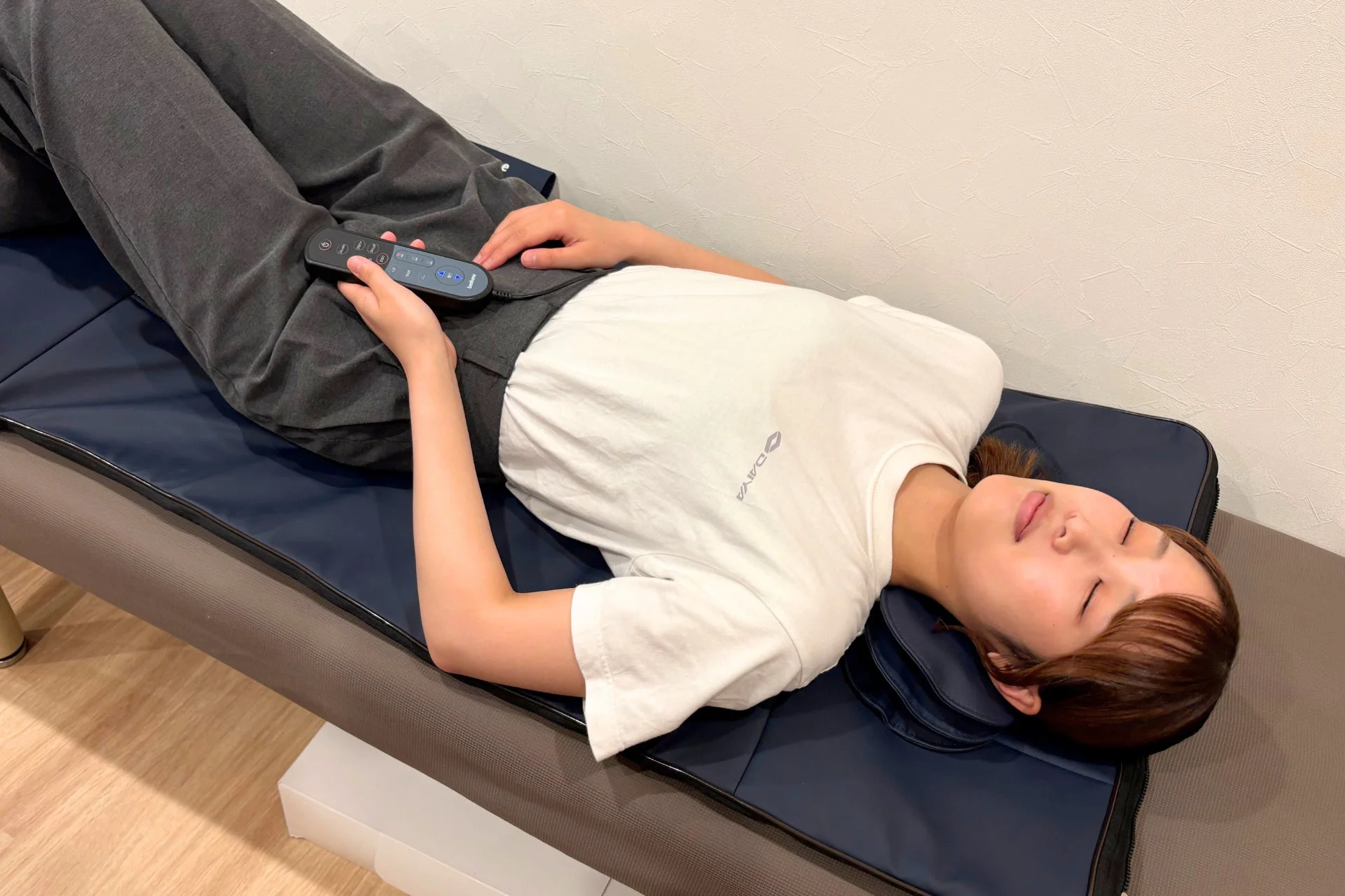 若い女性がリラックスした表情で、マッサージマットのようなものに横たわっています。手にリモコンを持ち、目を閉じて休息をとっている様子です。