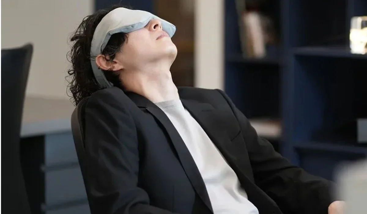アイマスクを着けて椅子に深くもたれかかり、仮眠をとっている男性の様子。オフィスらしき場所で仕事の合間に休憩しているように見えます。