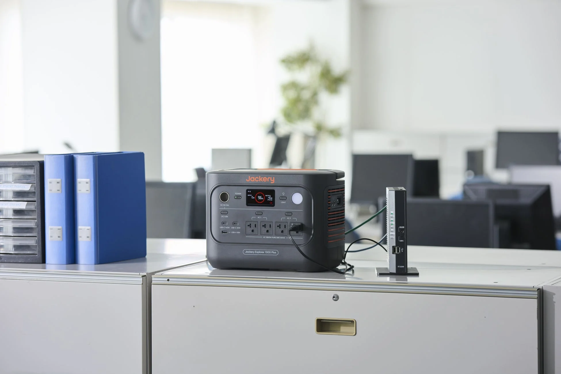 オフィス環境のデスクにJackery Explorer 1000 Plusポータブル電源が設置されており、別の機器に接続されています。