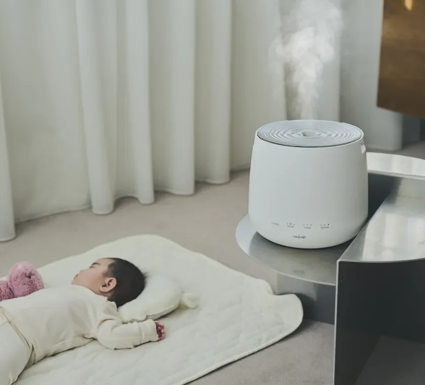 白い加湿器がミストを出し、その傍らで赤ちゃんが安らかに眠っています。清潔感のある空間で、赤ちゃんにとって快適な環境が整えられている様子です。
