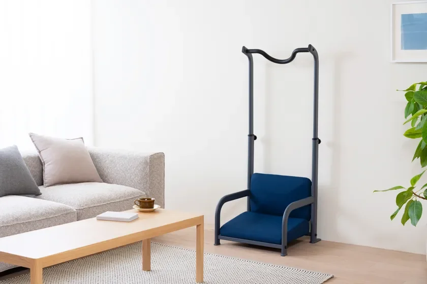 明るいリビングルームに、グレーのソファと木製のコーヒーテーブル、そして立ち上がりをサポートするような特徴的な青い椅子が配置されています。