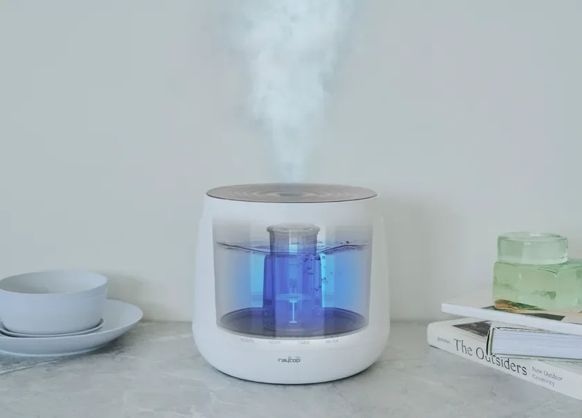 白いモダンな加湿器がミストを噴出し、内部の青い光が特徴的です。テーブルの上に置かれ、周囲には食器や本が配置されています。