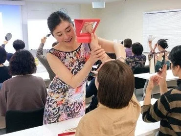 女性の講師が、赤い三角形の器具を使って別の女性に美容または姿勢に関するデモンストレーションを行っています。