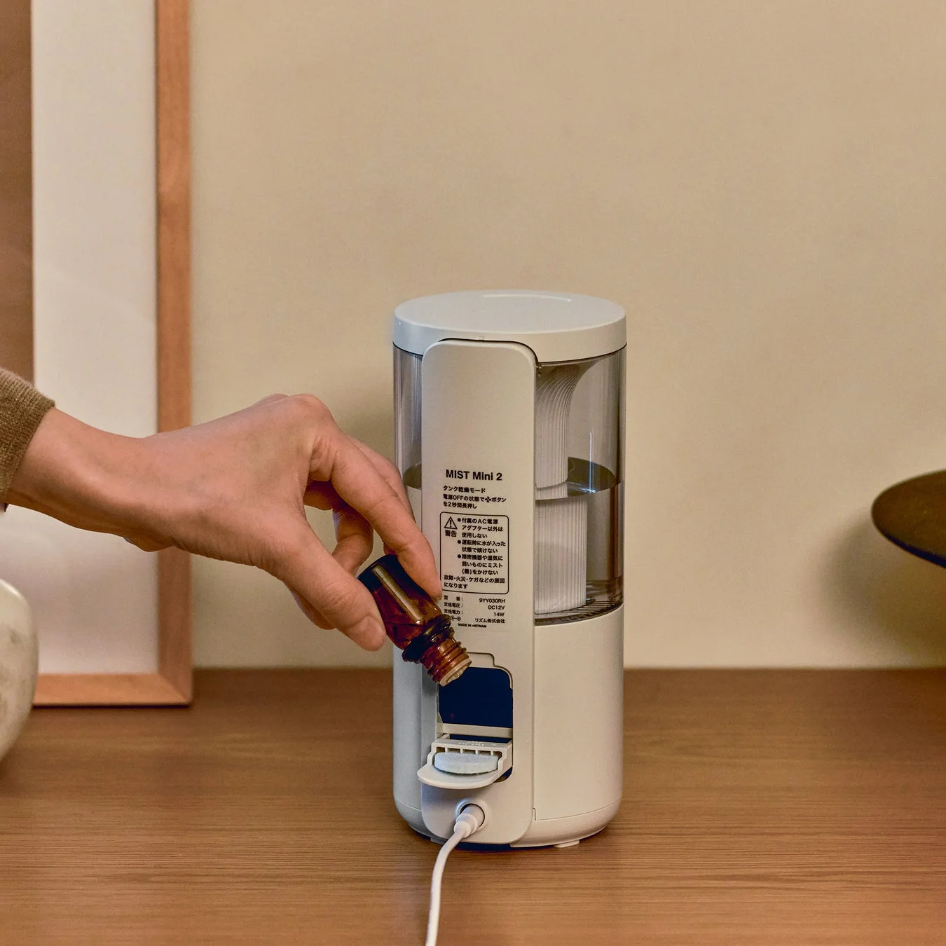 木製のテーブルに置かれた白いコンパクトなディフューザーに、手がエッセンシャルオイルを注いでいる様子
