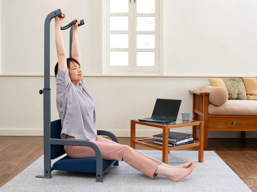 明るい部屋で女性が家庭用運動器具に座り、両腕を上へ伸ばしてストレッチをしている。